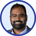 A man with glasses and a beard smiles, wearing a grey blazer and white shirt against a light blue background, epitomizing the spirit of advancing technology.