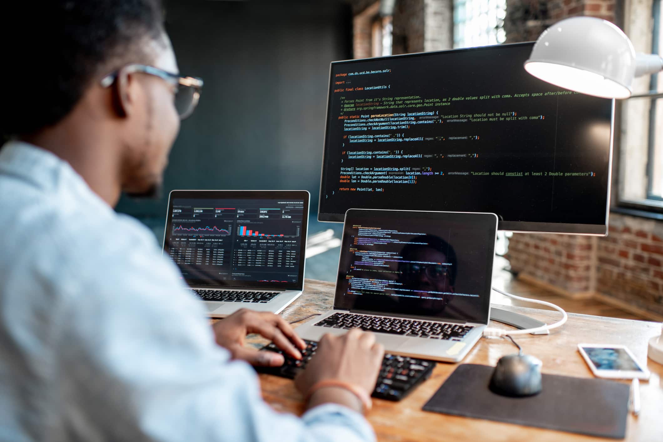 A person sits at a desk working on three screens displaying code, graphs, and emails in a modern office setting, meticulously reviewing the cybersecurity checklist.