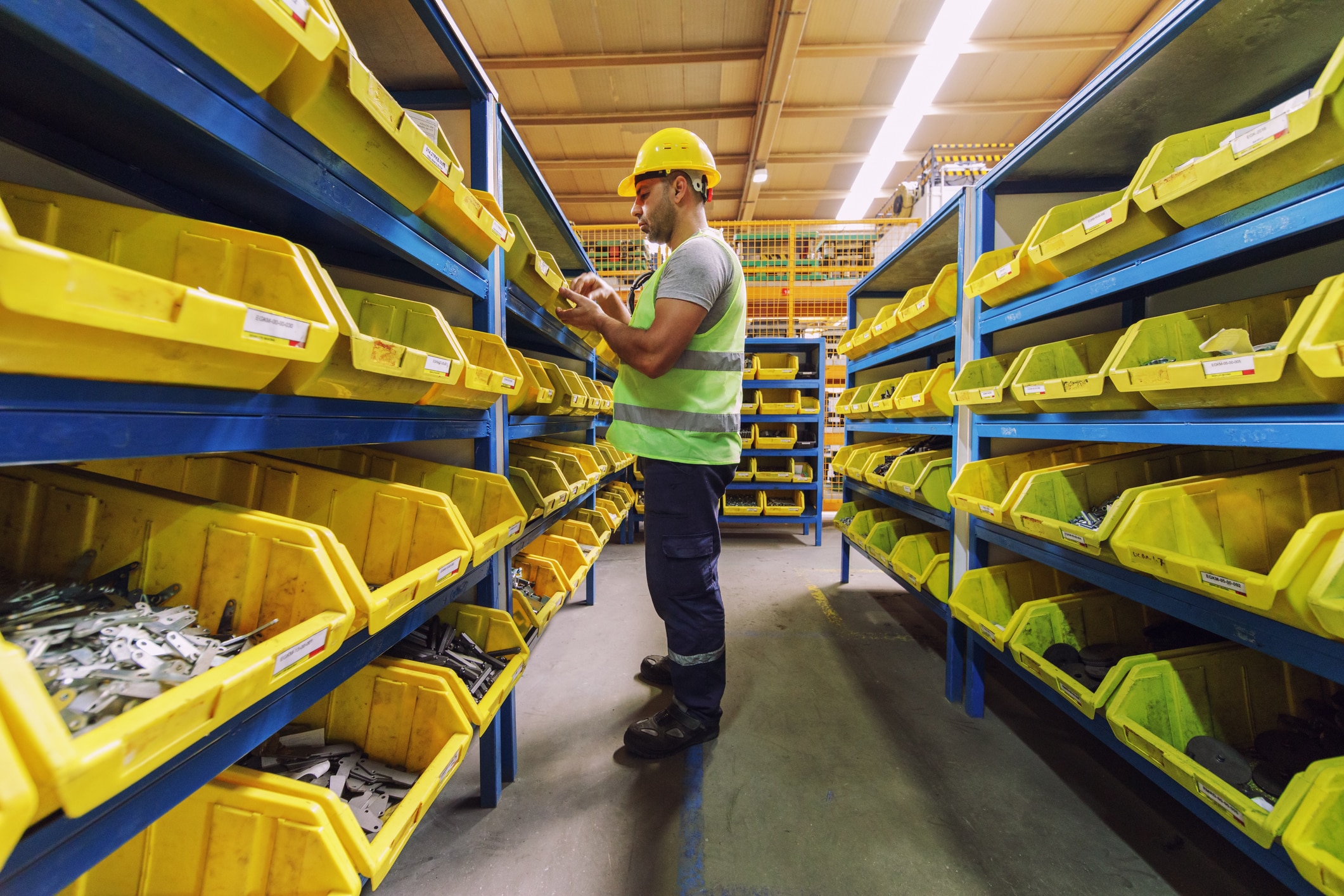 A worker wearing a hard hat and reflective vest organizes inventory in yellow bins on shelves in a warehouse, showcasing storeroom management best practices.