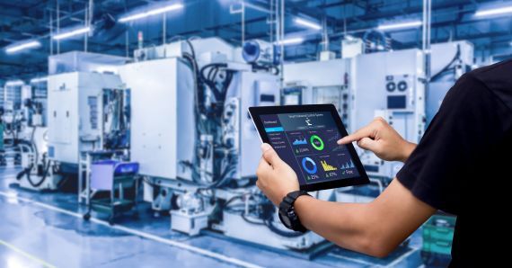 Person holding a tablet with data analytics interface in a factory, monitoring production efficiency amidst industrial machinery in the background.