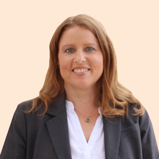 Allison Kuhn, with long, light brown hair, smiles warmly in a white blouse and gray blazer, set against a beige background.