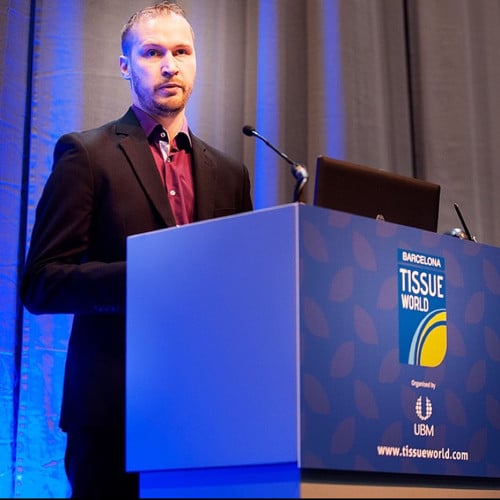 A person in a suit stands at a podium with a "Tissue World" sign, giving an insightful presentation on Insight Driven Manufacturing in a professional setting, aiming to transform your business.